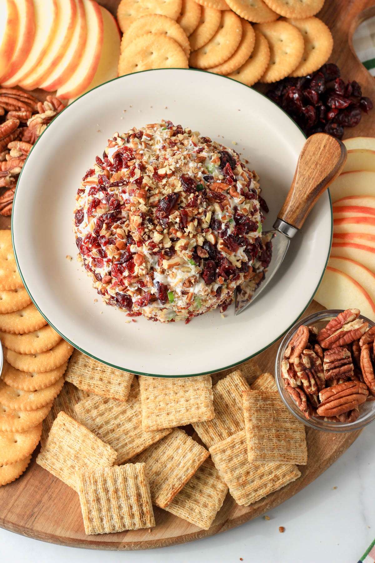 A top down picture of the vegan holiday cheese ball on a white plate with a cheese knife to the right surrounded by crackers, nuts, and apples.