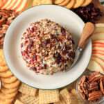 A close up of a vegan holiday cheese ball topped with pecans and dried craisins on a white plate with a green rim and a cheese knife to the right.