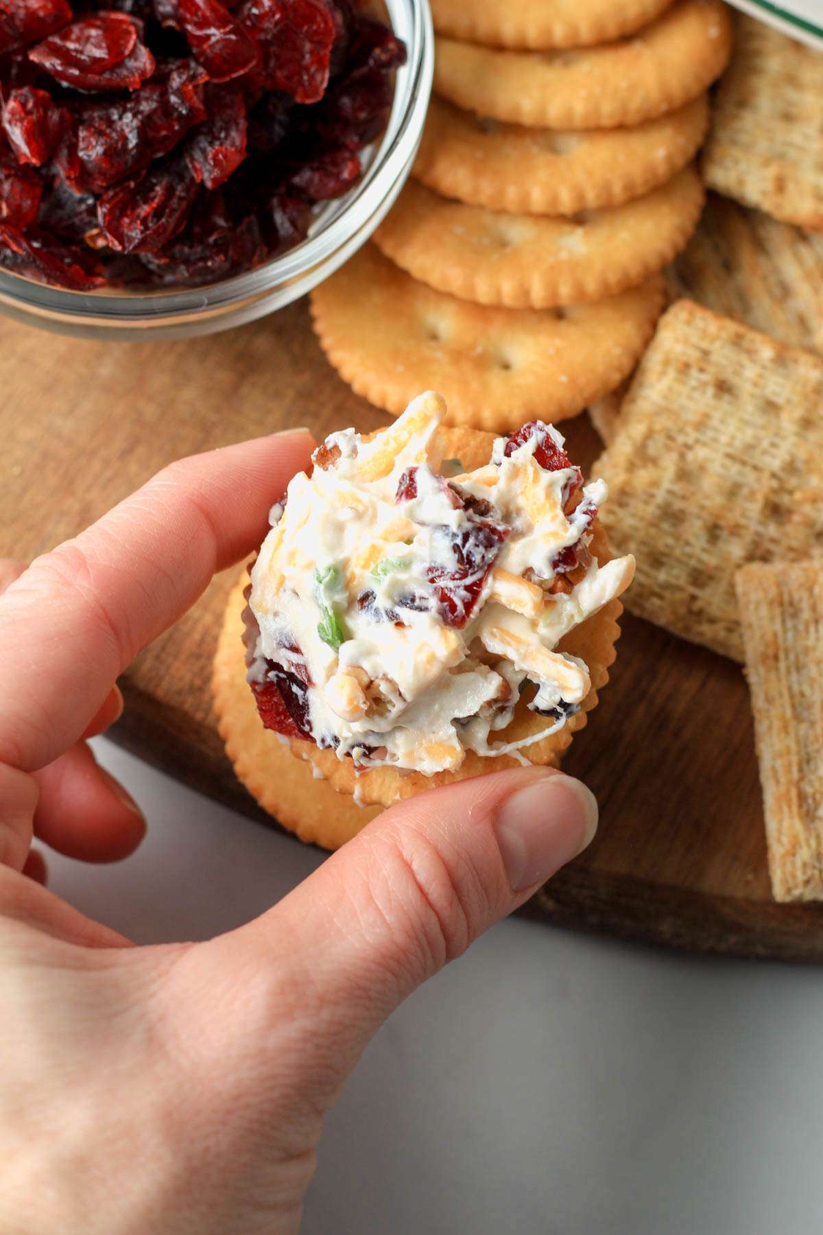 A hand holding a cracker topped with pecan and cranberry vegan cheese ball.