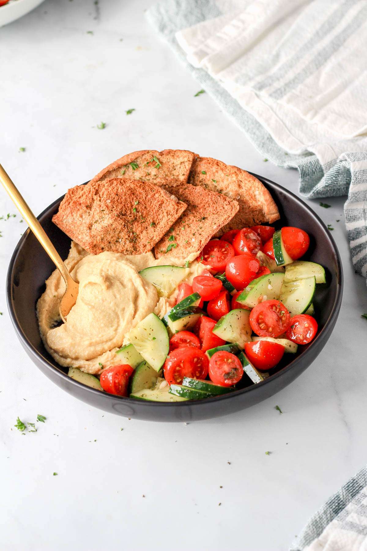 A black bowl filled with hummus, veggies, and pita chips on a white counter.