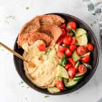 A close up of a black bowl filled with hummus, pita chips, and a cucumber and tomato salad with a gold spoon in the left side of the bowl.