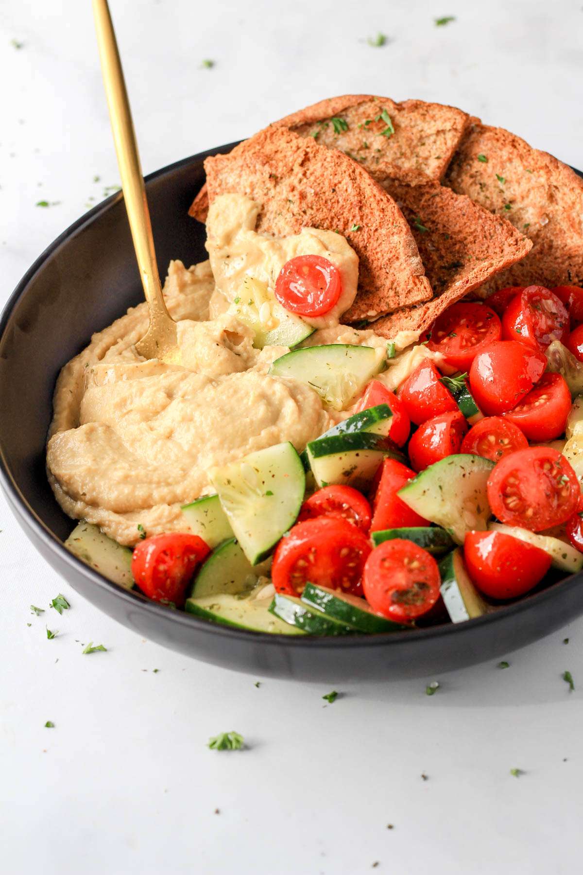 A black bowl with a healthy vegan hummus bowls blend of hummus, vegetables, and pita chips on a white counter.
