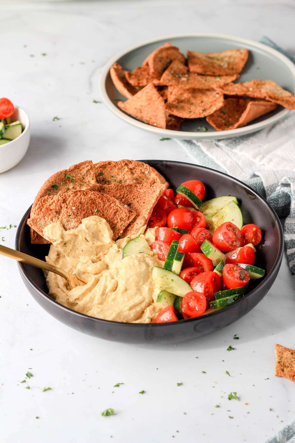 Healthy vegan hummus bowls from the side with a spoon and a plate of pita chips in the back.