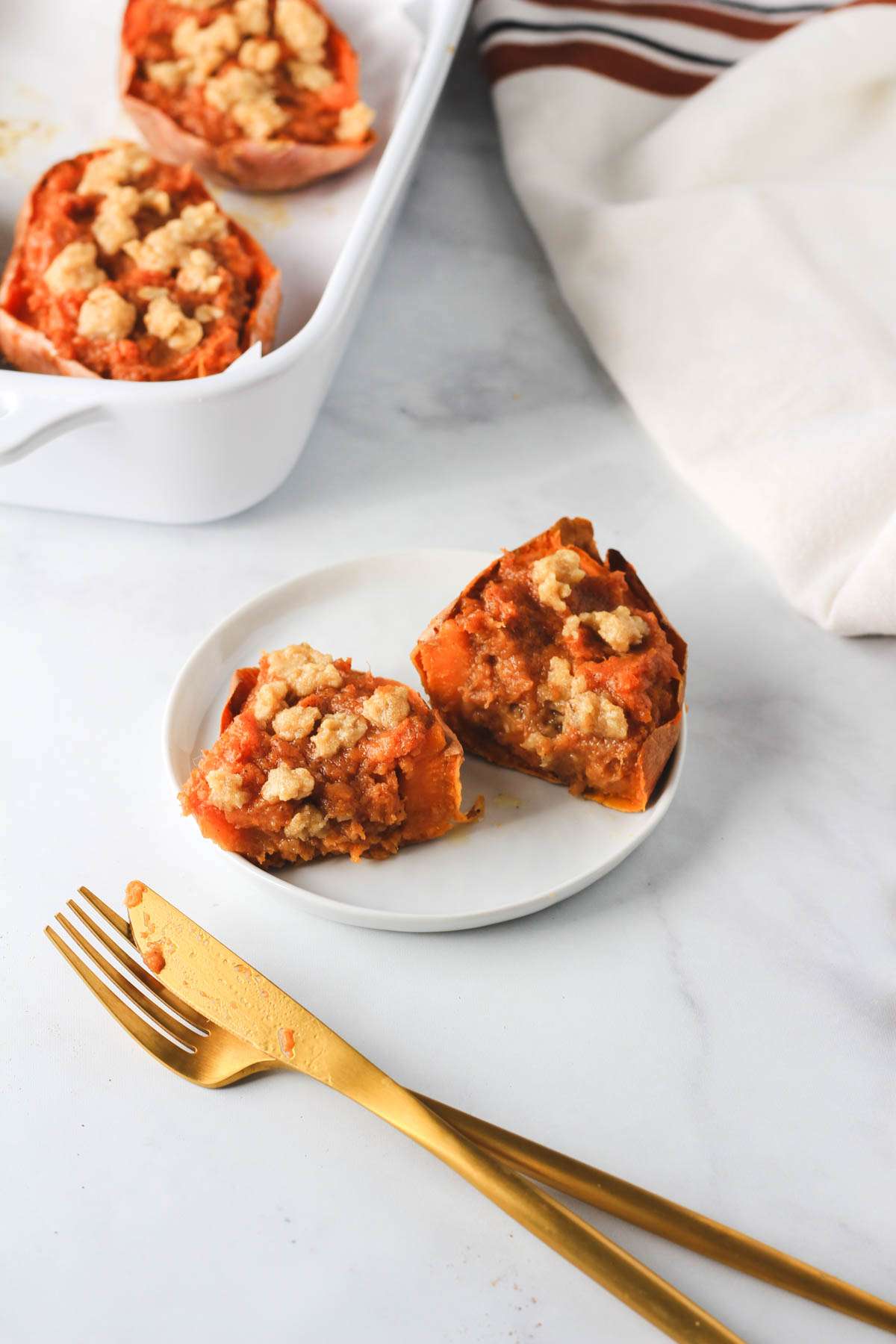 A small white plate with a dairy-free twice baked sweet potato after being cut in half with cutlery in the bottom left and a baking dish in the top left.