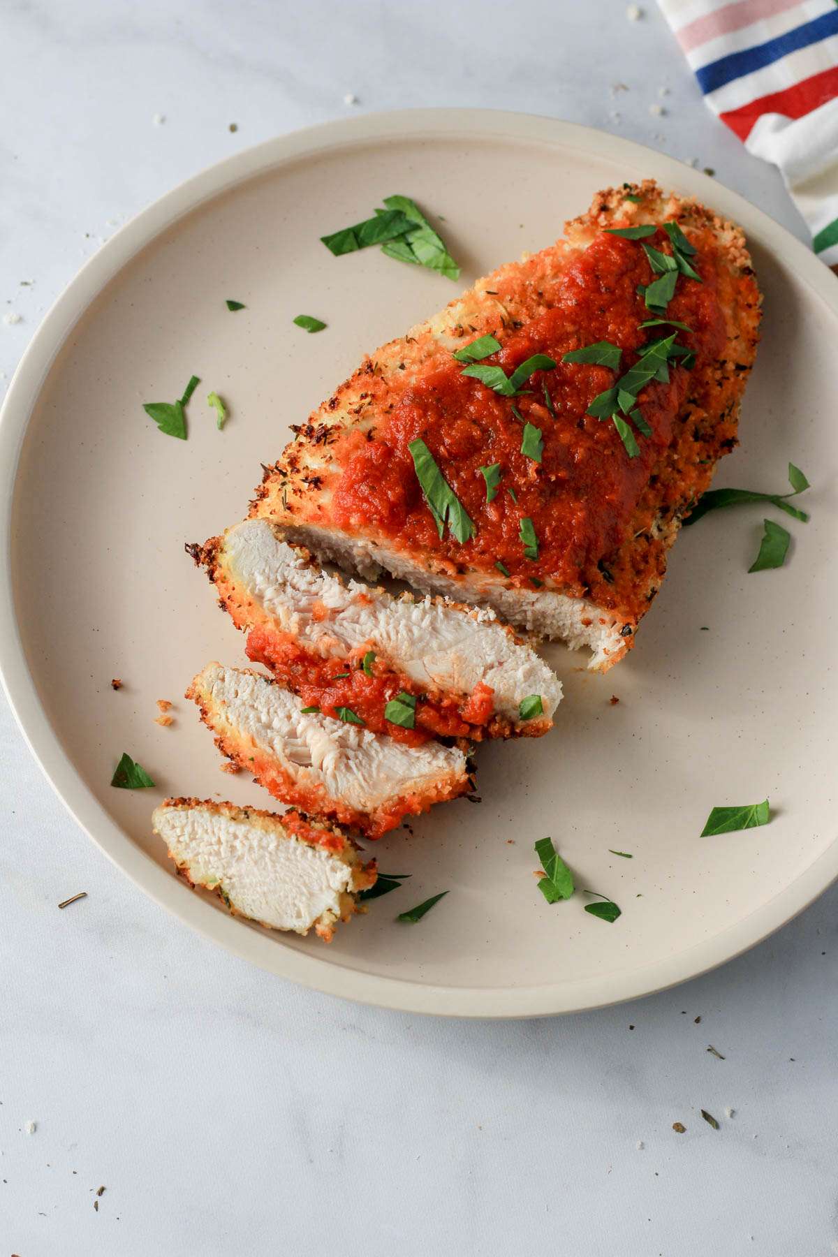 A white plate with dairy-free air fryer chicken parmesan sliced and topped with fresh parsley on a white counter.