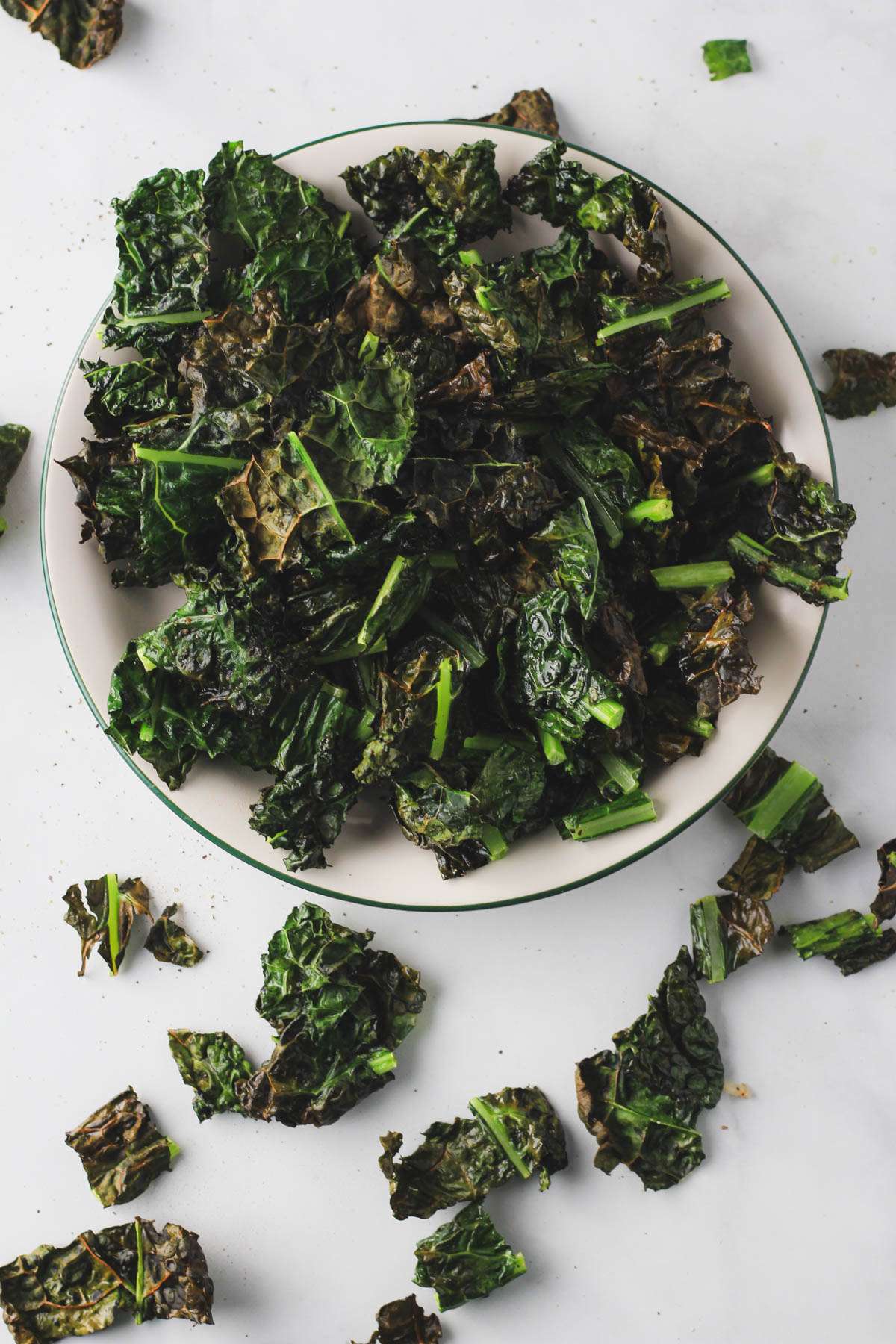 Vegan air fryer kale chips on a white plate with a green rim on a white counter with a few kale chips scattered on the counter.