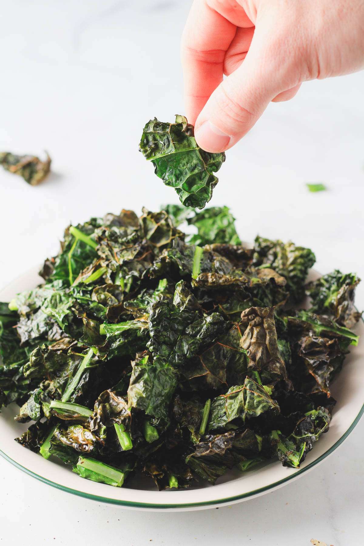 A hand holding one air fryer kale chip over a white plate topped with vegan kale chips.