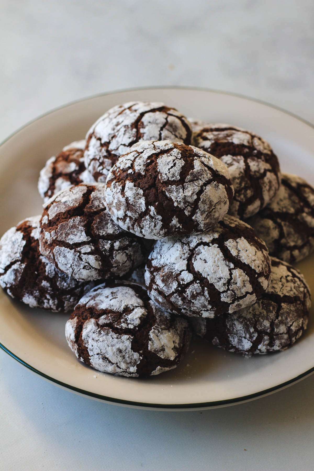 A pile of powdered sugar coated chocolate crinkle cookies on a white plate with a green rim.