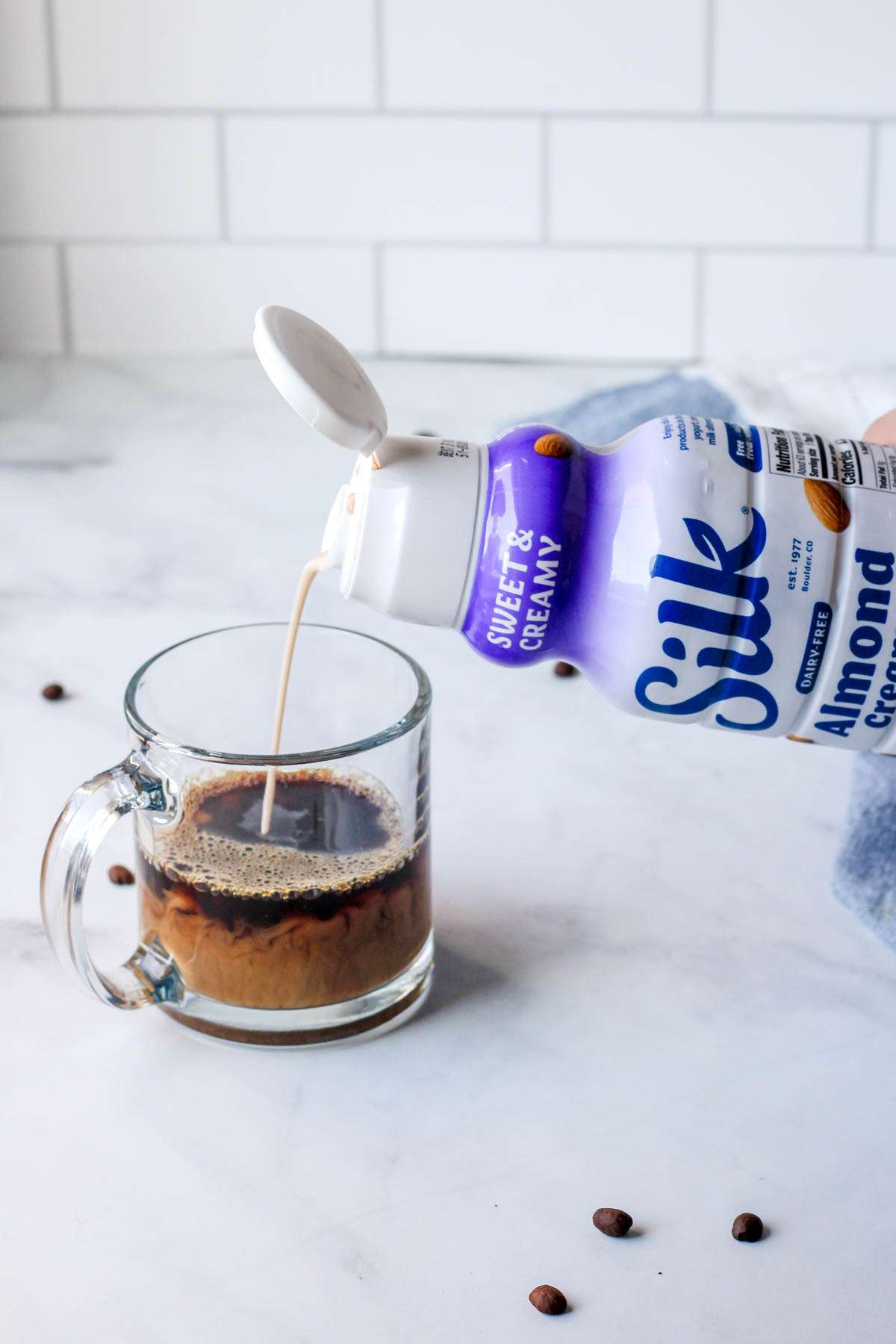 A bottle of Silk coffee creamer being poured into a cup of coffee on a white counter.