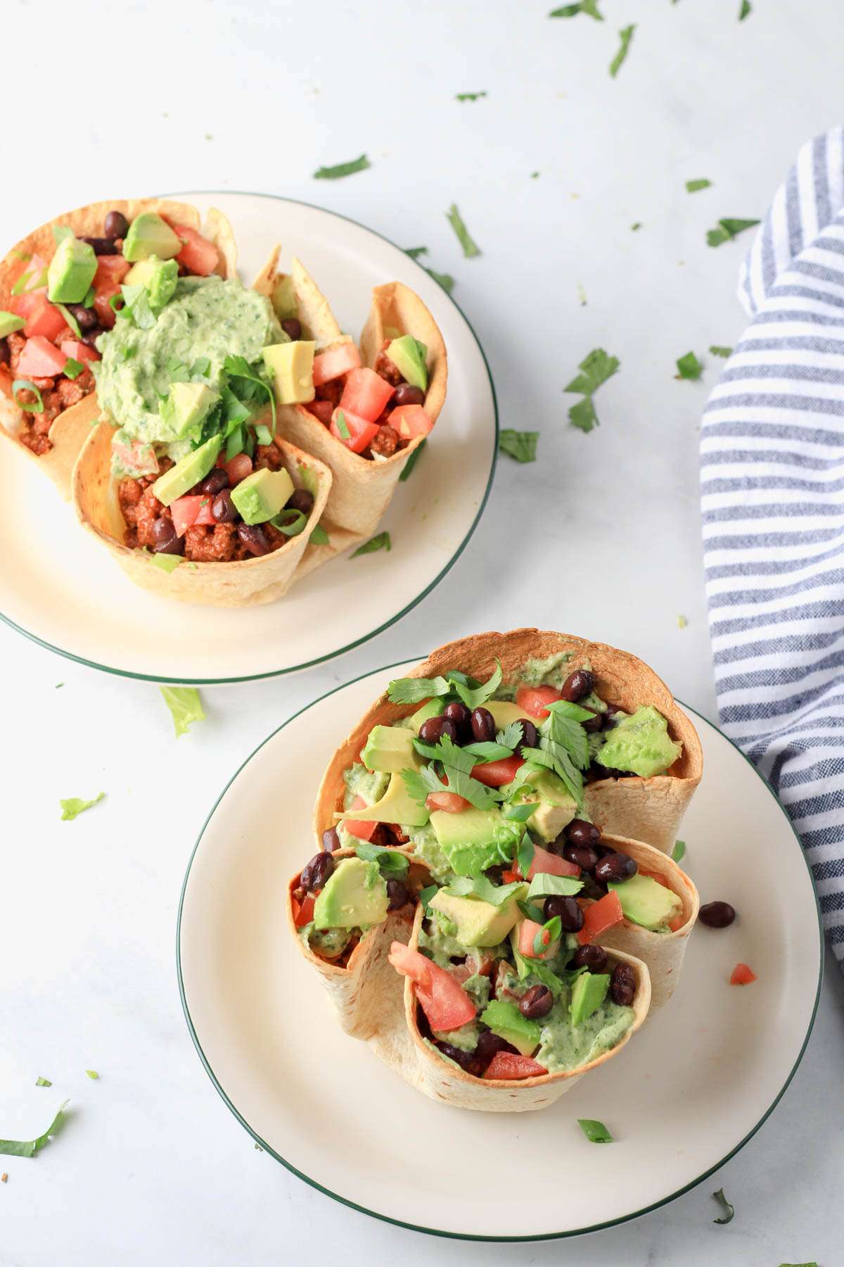 Two white plates with green rims on a white counter with taco salad in a baked flour tortilla bowl.