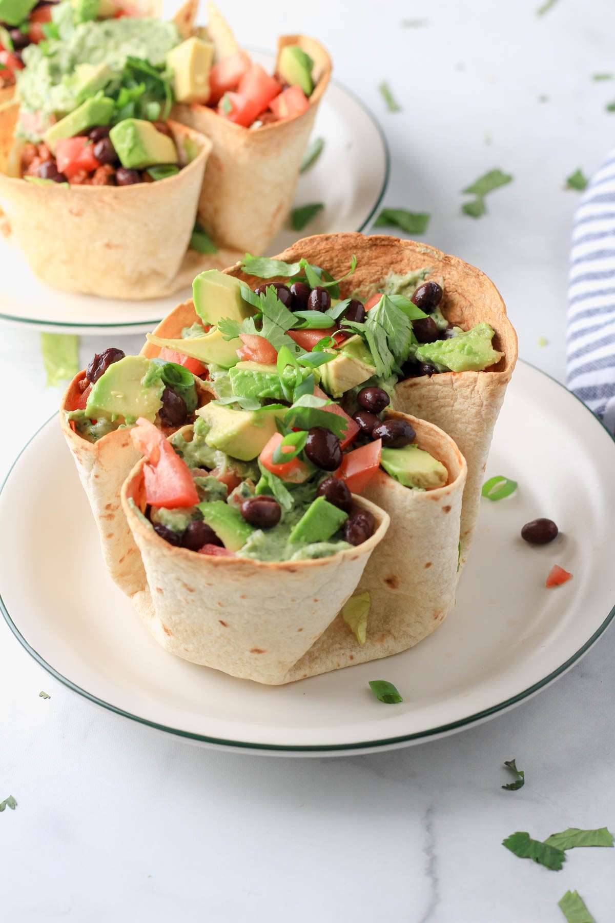 A baked flour tortilla bowl on a white plate filled with lettuce, taco meat, beans, avocado, tomato, cilantro, green onion, and dressing.