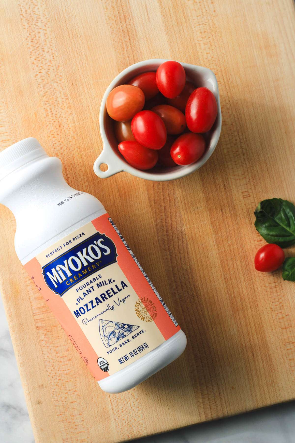 A wooden cutting board with a bottle of liquid mozzarella in the bottom left and a small bowl of grape tomatoes in the top.