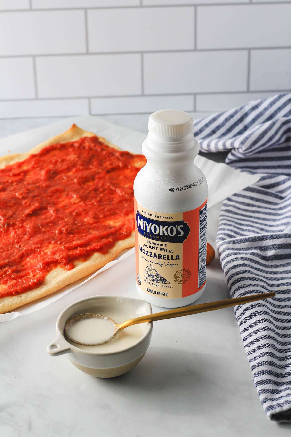 A flatbread pizza in the back left with a bottle and bowl of miyoko's plant cheese in front.