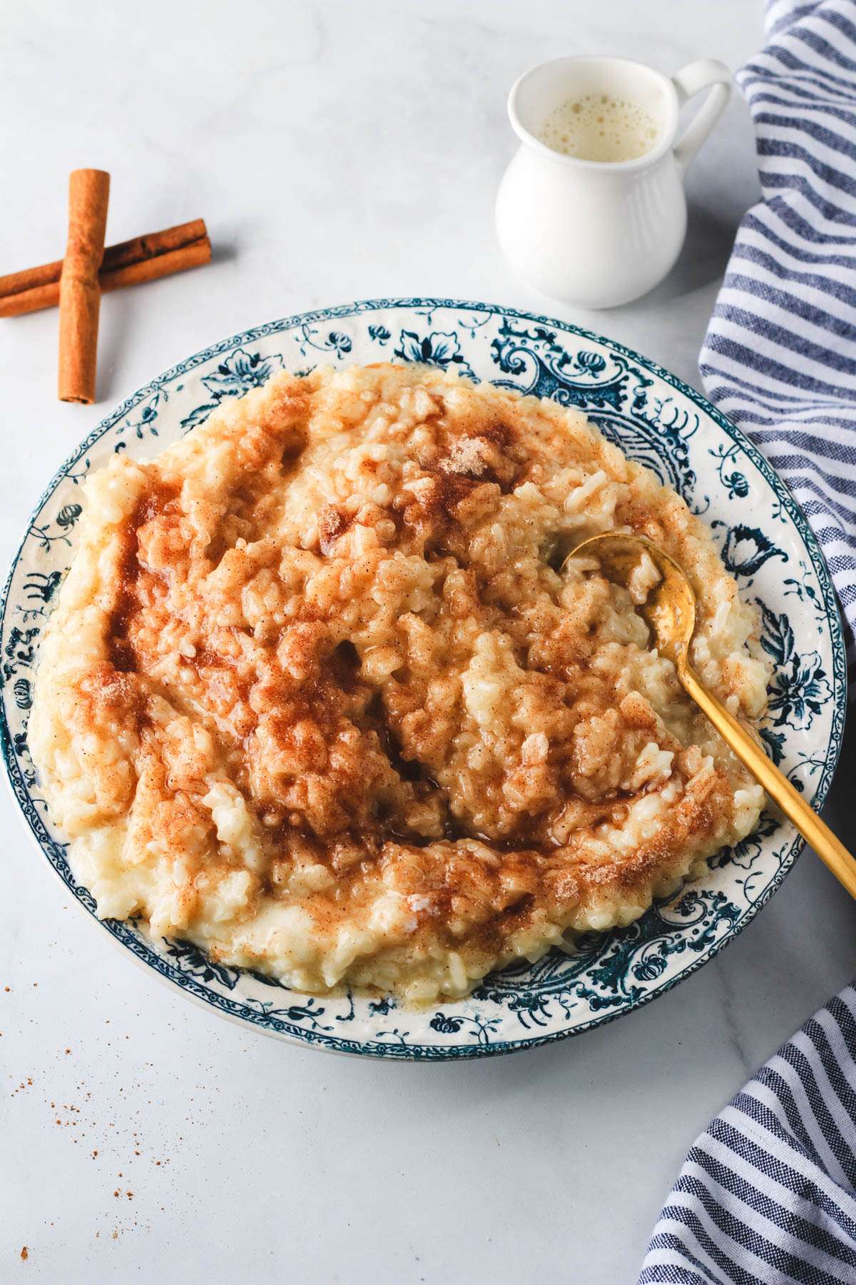 Vegan oat milk rice pudding in a blue and white rimmed bowl with a gold spoon on a white counter.