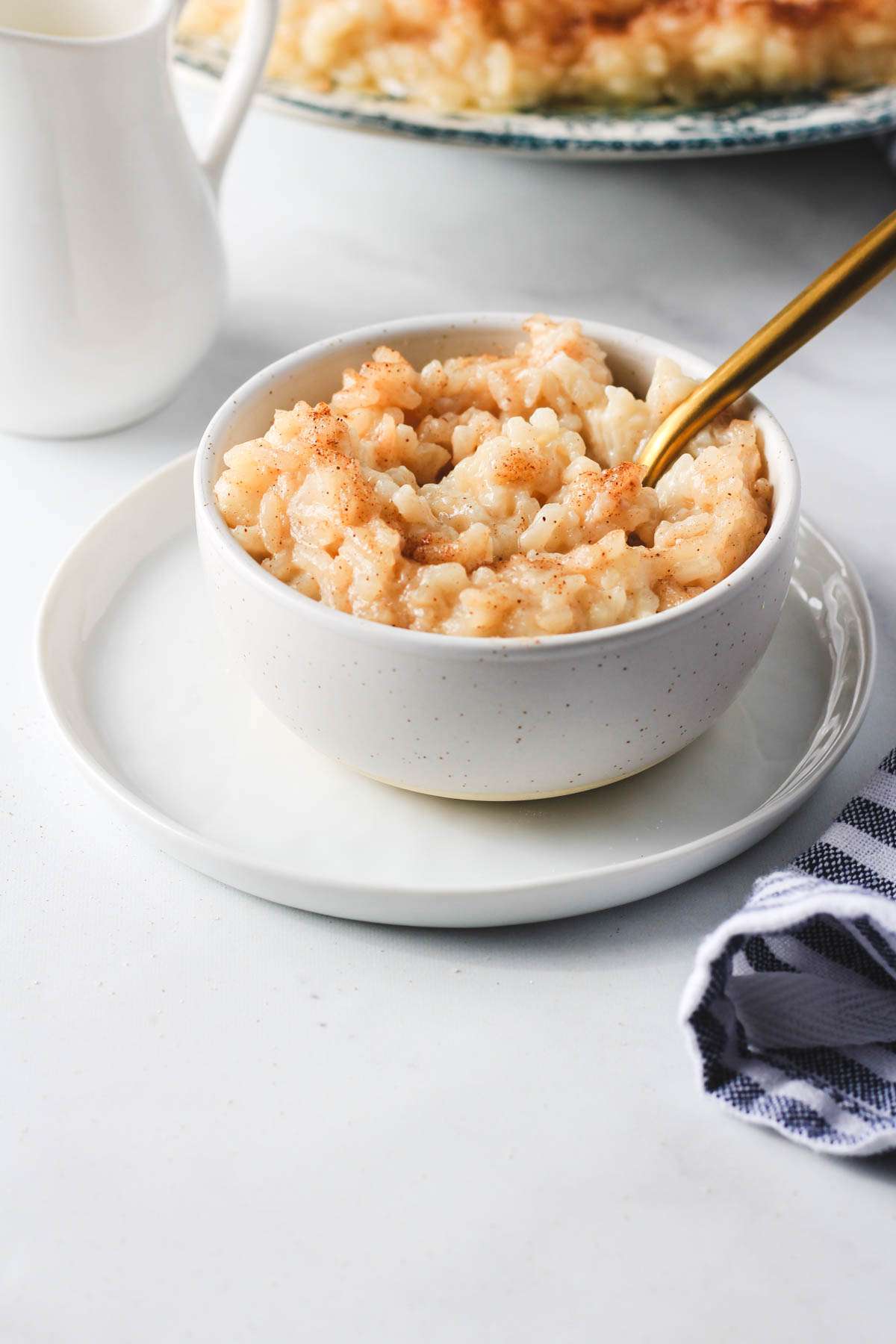 A white bowl with oat milk rice pudding on top of a white plate with a gold spoon sticking out of the bowl.