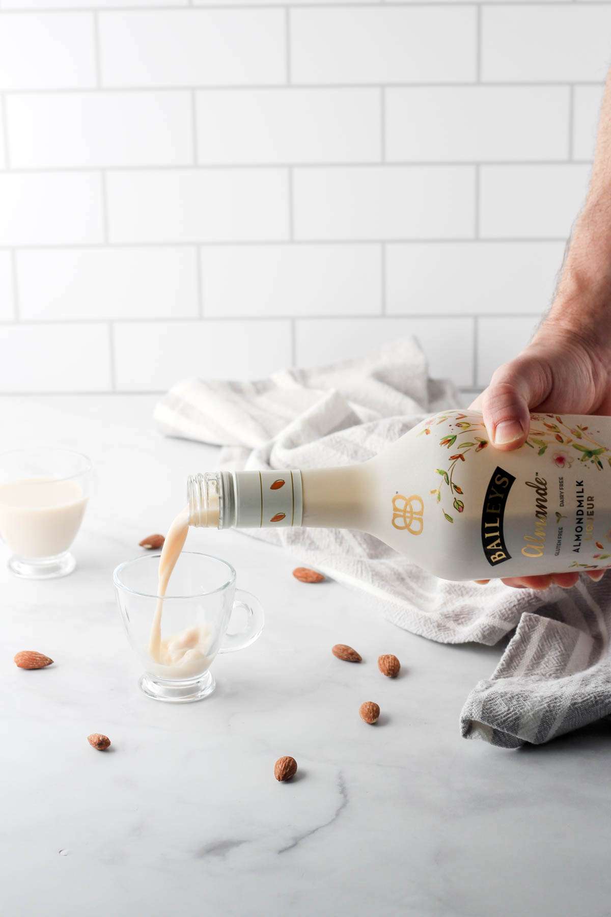 A hand pouring a glass of Bailey's Almande into a small clear glass cup.