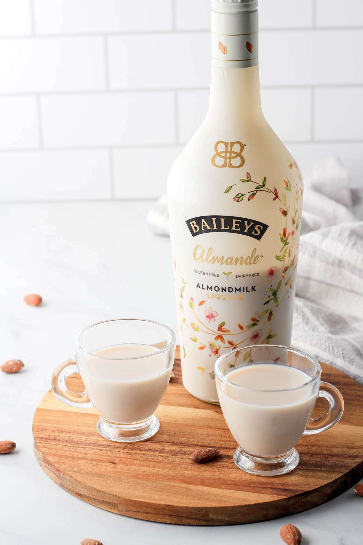 A bottle of Bailey's Almande for this review on a wooden tray with two glasses of almond milk liquor in the front.