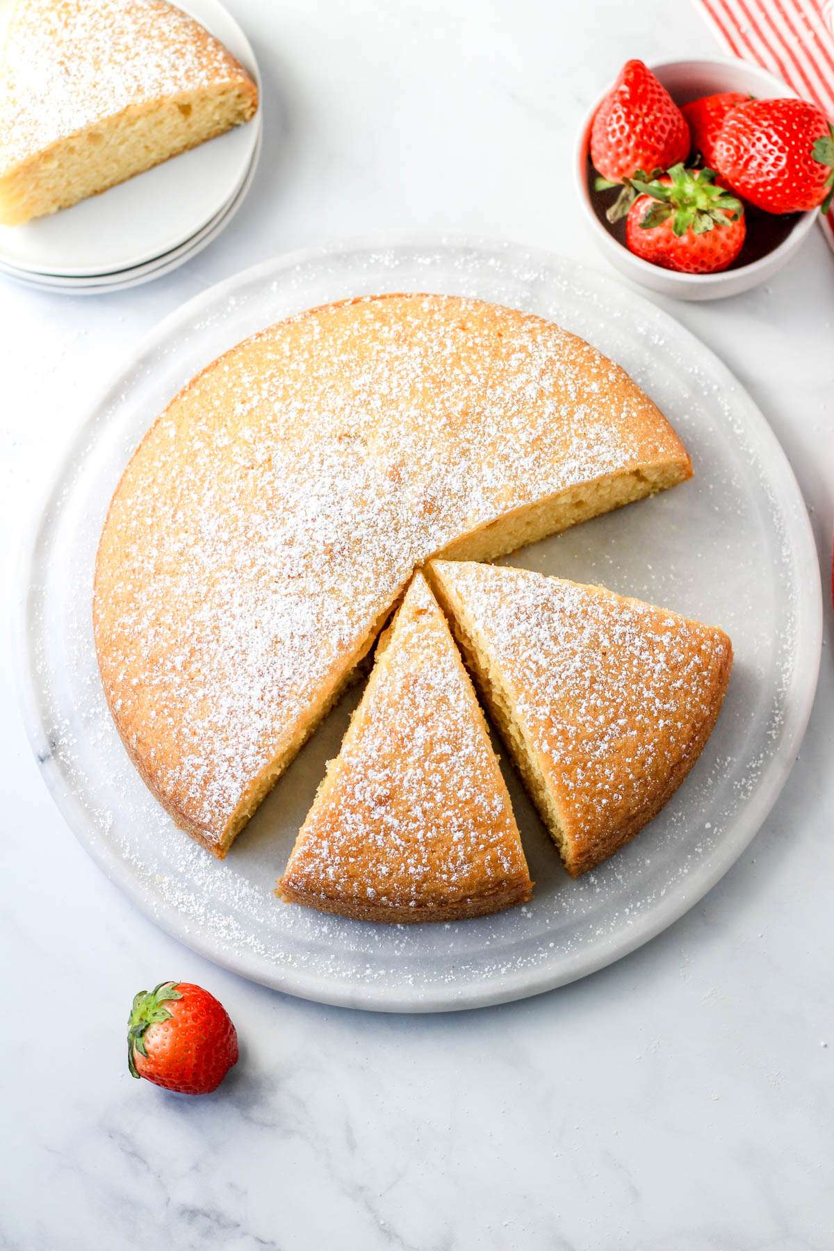 A dairy-free yogurt cake cut into slices on a marble cake stand with strawberries in the top right and bottom left.