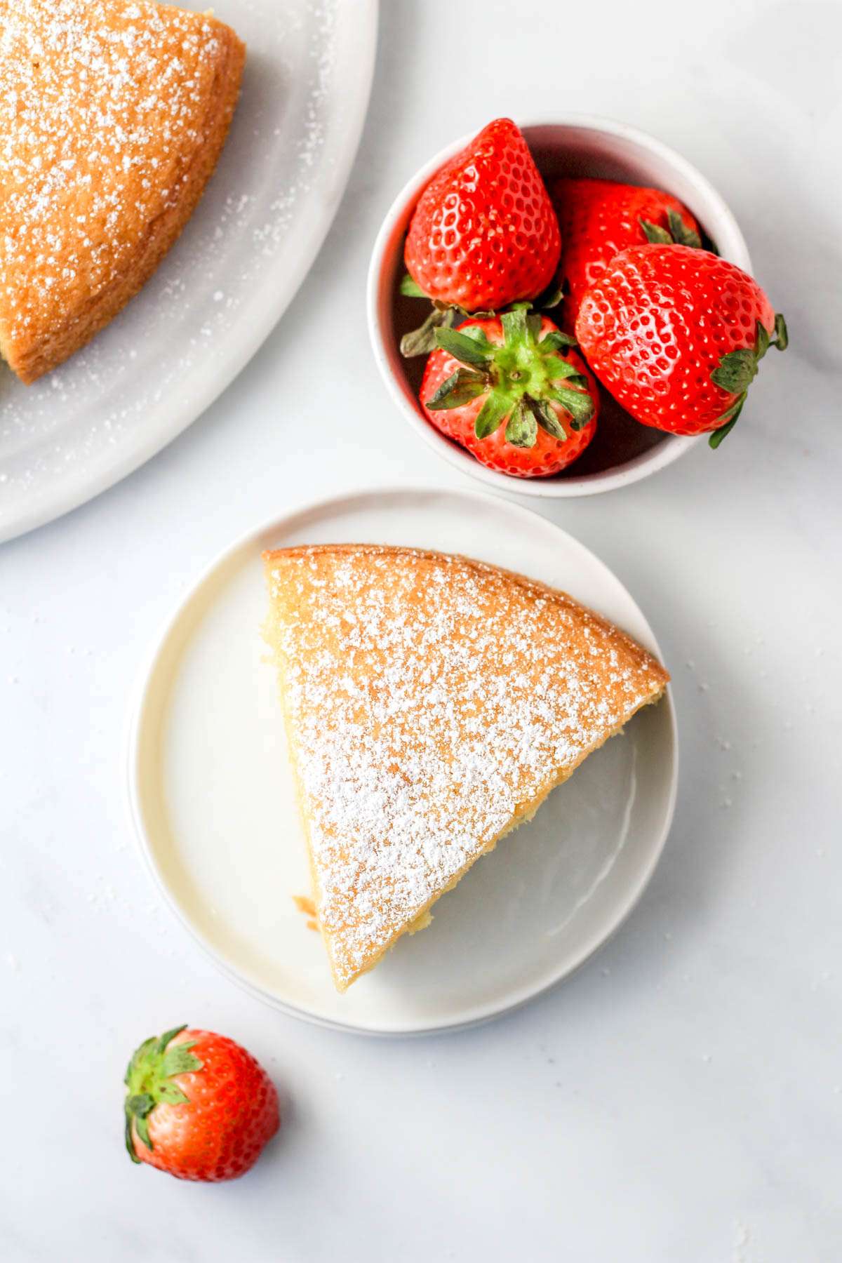 A top down photo of a slice of yogurt cake dusted with powdered sugar on a white plate with strawberries in the top left and bottom right.
