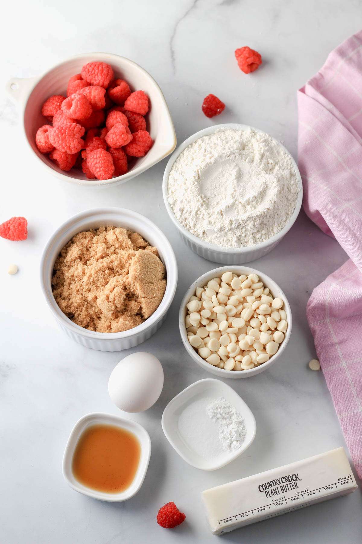 Ingredients for raspberry white chocolate chip cookies on a white counter with a pink dish towel to the right.