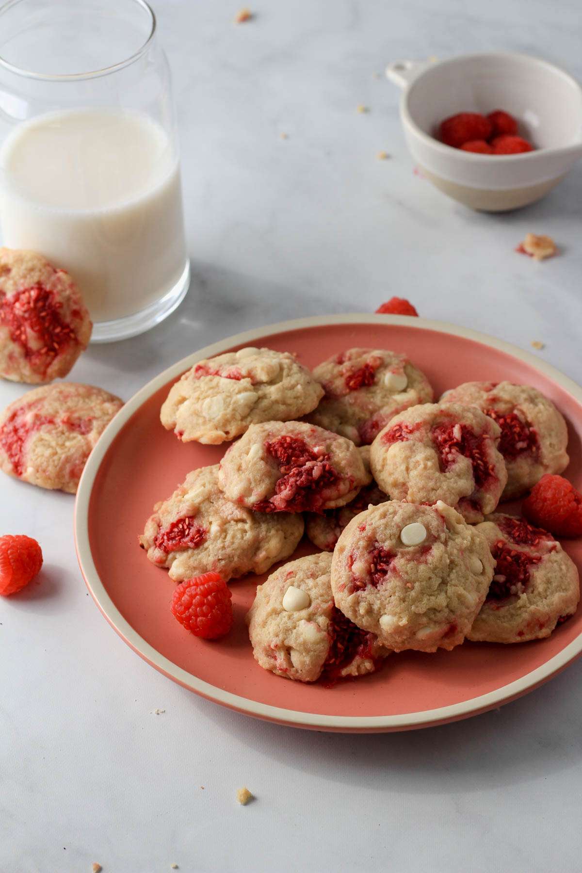 A pink plate topped with a few raspberry white chocolate chip cookies with a glass of oat milk in the back left.