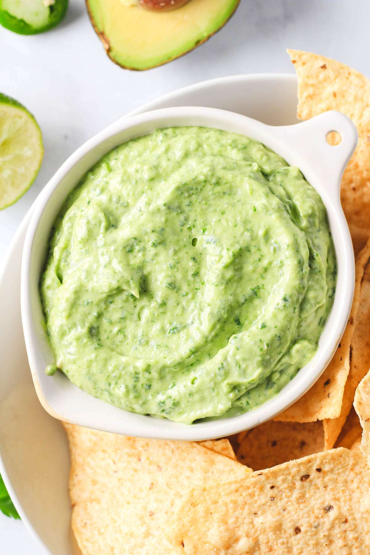 A top down photo of a small white bowl filled with vegan cilantro avocado crema in a larger bowl of chips.