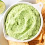 A close up of a white bowl of vegan avocado cilantro lime crema surrounded by tortilla chips.