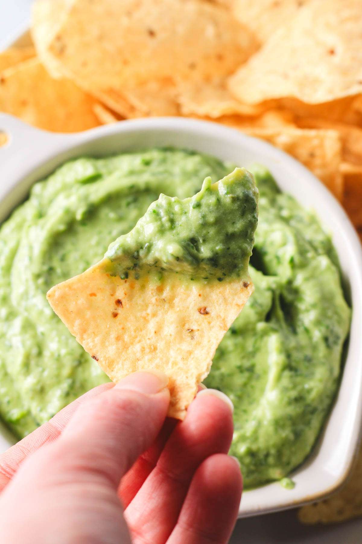 A hand holding a tortilla chip dipped in the creamy vegan avocado dip.
