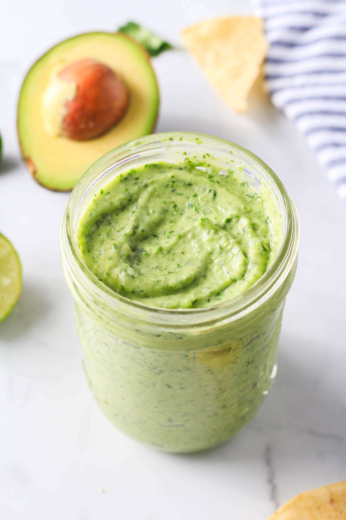 A glass mason jar on a white counter filled with vegan avocado dip with a white and blue towel to the right.