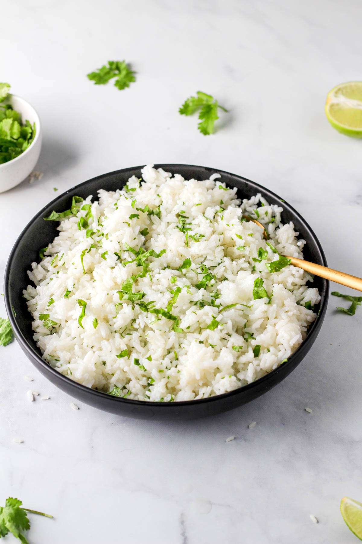 Dairy-free cilantro lime rice in a black bowl with a gold spoon on a white counter with additional cilantro and lime around.