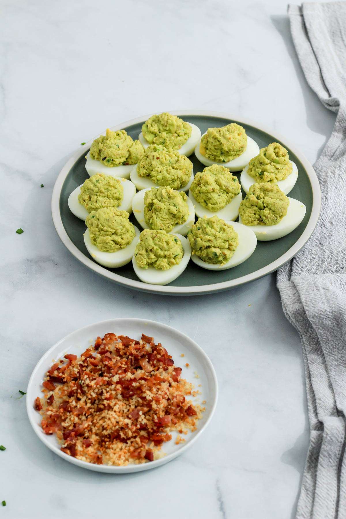 No mayo deviled eggs with avocado on a green plate in the back right with a small white plate of bacon and panko breadcrumbs on a white plate in the front left.