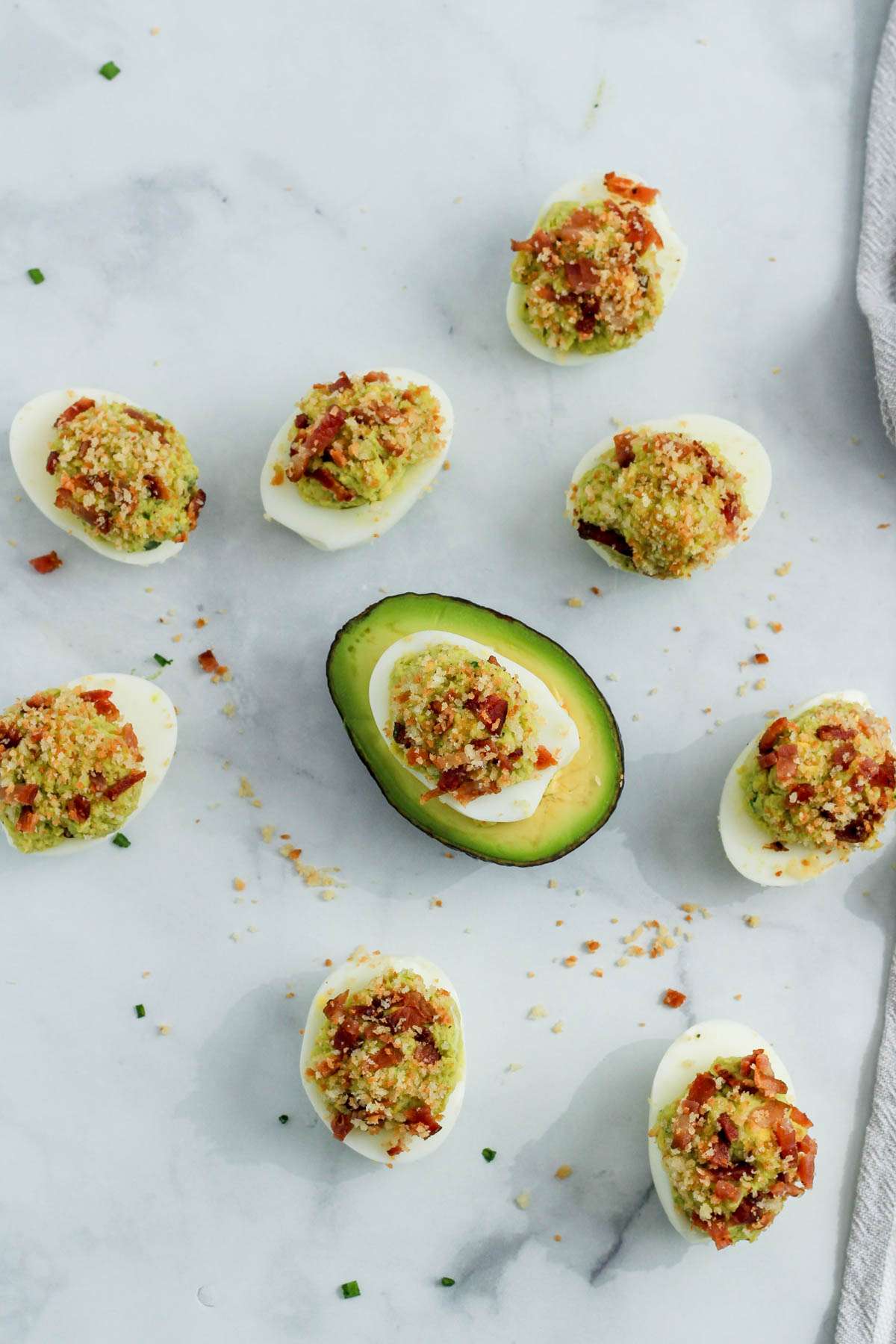 A scattering of avocado deviled eggs on a white counter topped with bacon and toasted breadcrumbs.