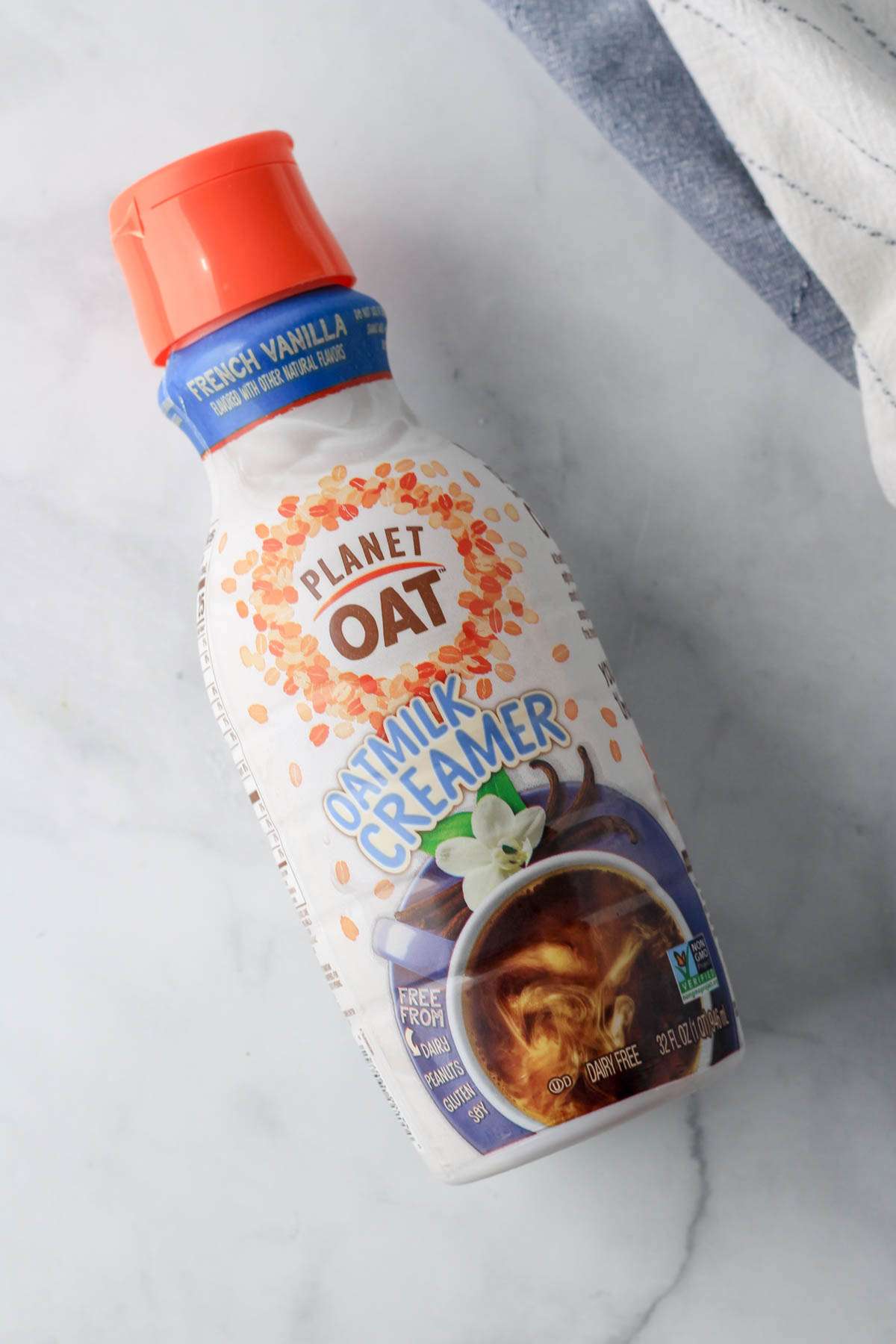 A large bottle of dairy-free, planet oat coffee creamer on a white counter with a blue and white dish towel to the right.