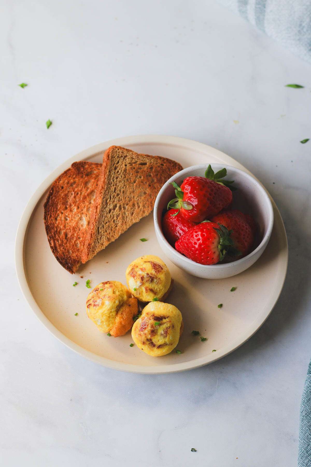 A top down picture of a cream plate topped with three dairy-free egg bites, a small side of toast, and a small bowl of fresh strawberries.