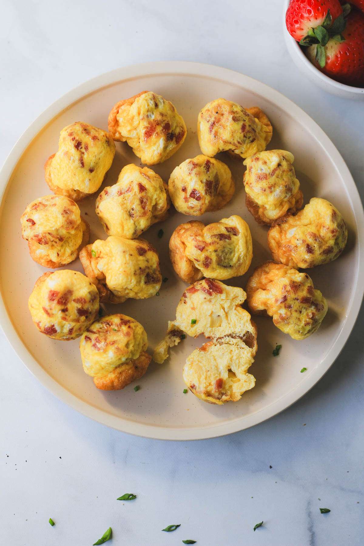 A cream plate covered in dairy-free air fryer egg bites with one egg bite cut down the middle and a small bowl of strawberries in the top right corner.