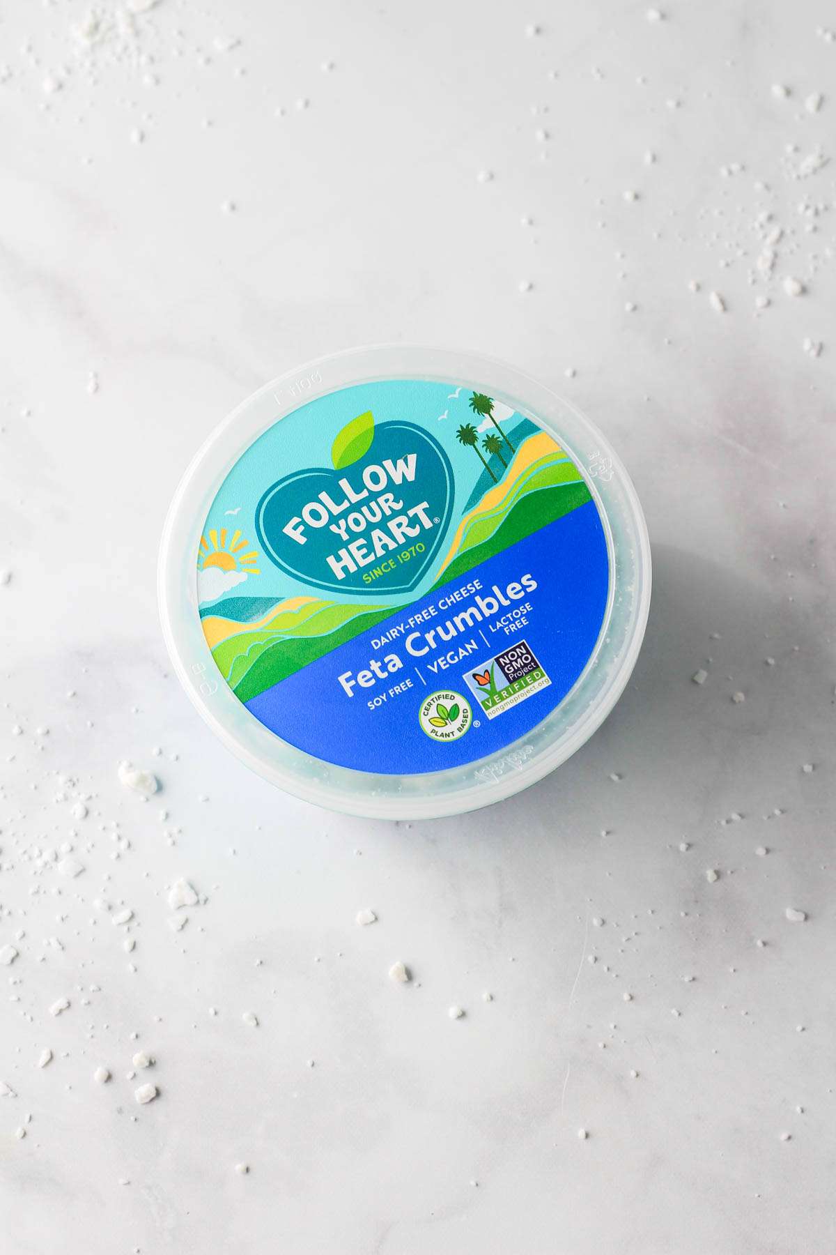 A blue and green container of Follow Your Heart Feta Cheese Crumbles on a white counter.