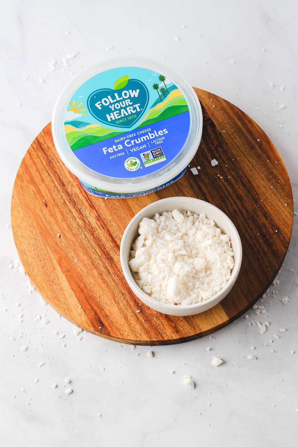 A round wooden plate topped with a small white bowl of vegan cheese and a small container of follow your heart feta cheese crumbles.