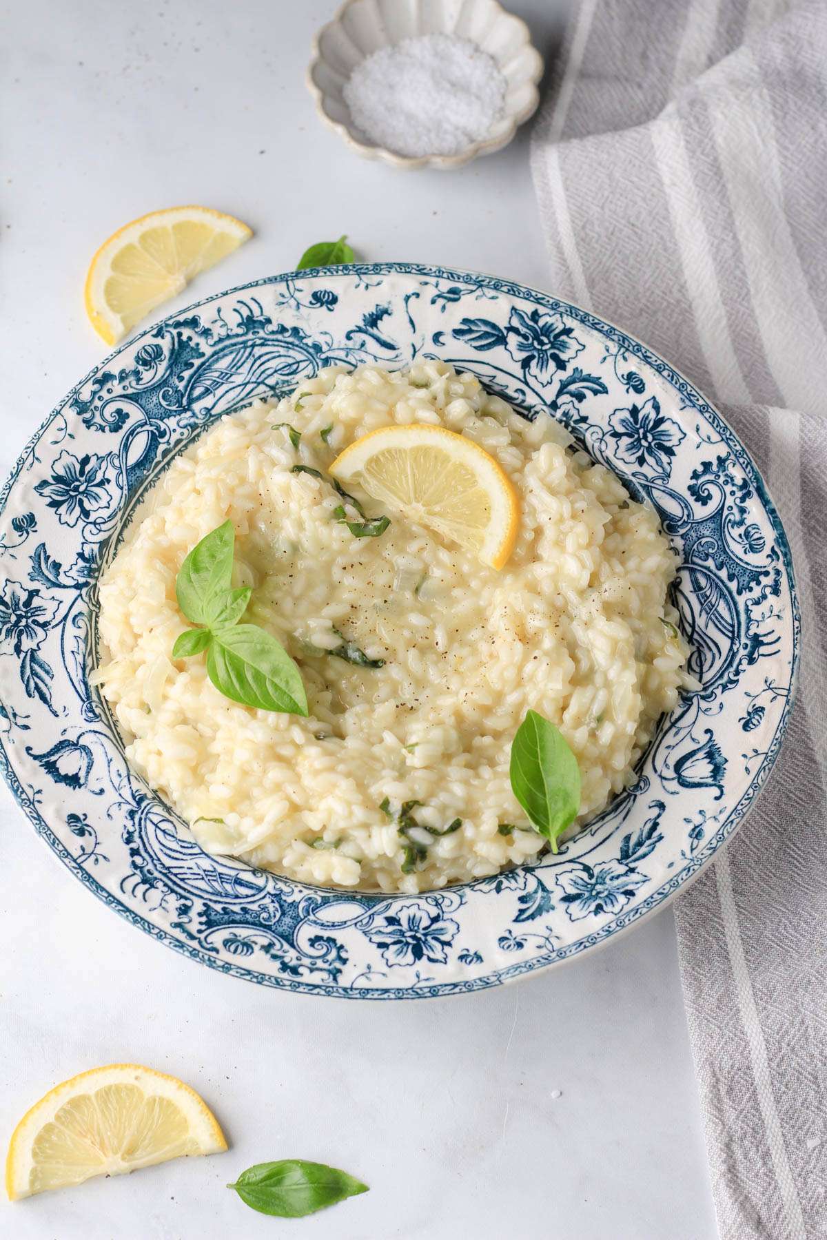 A white and blue bowl filled with vegan lemon basil risotto on a white counter with a small pinch bowl of salt in the back and a grey and white dish towel to the right.