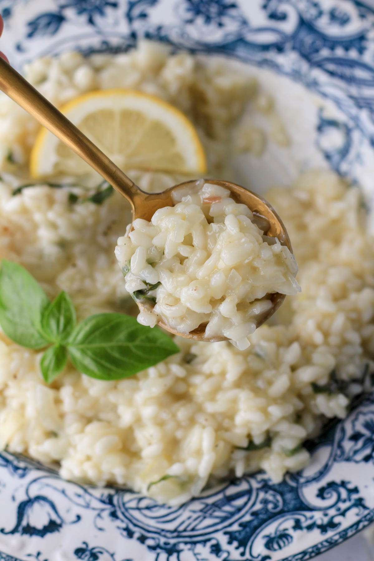 A gold spoon with a scoop of vegan lemon basil risotto over a blue and white rimmed bowl of risotto.