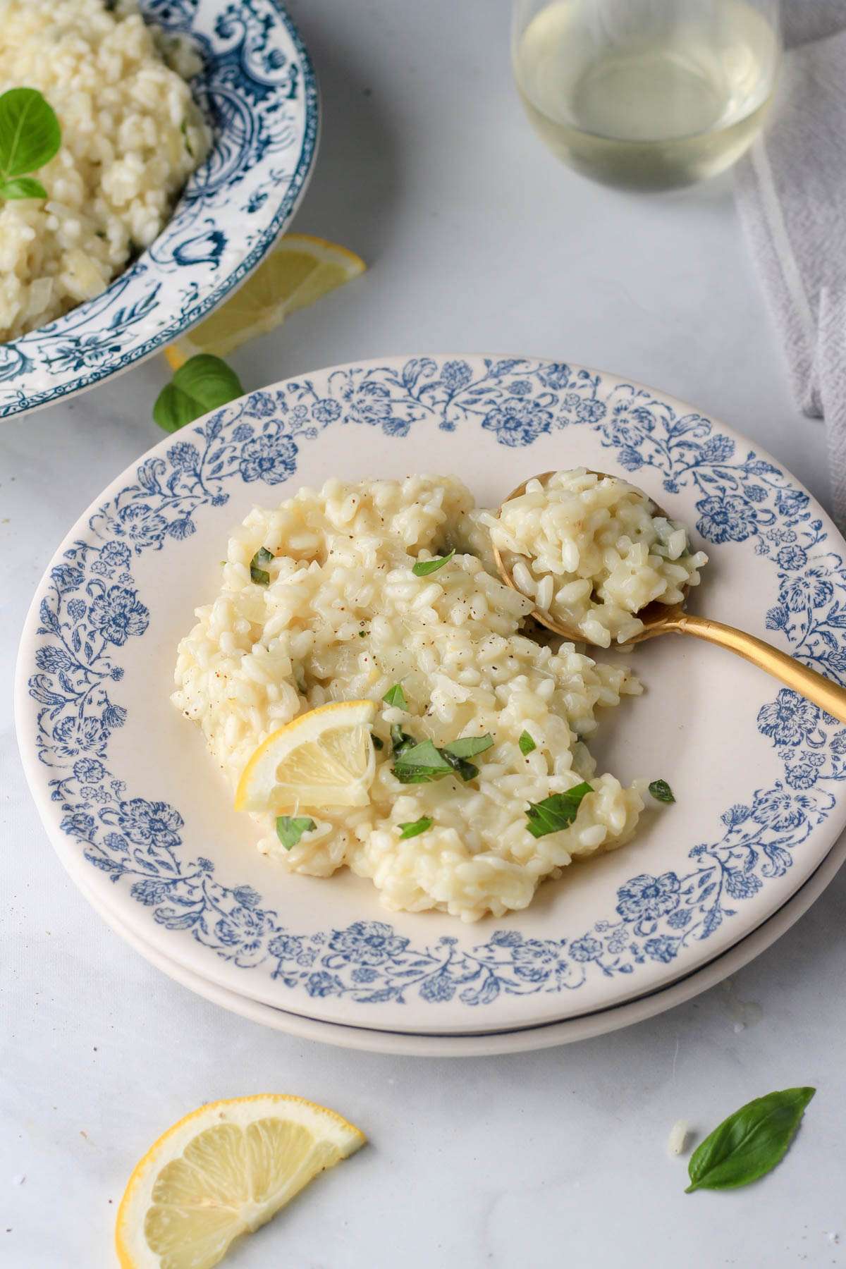 A stack of plates with a scoop of vegan lemon basil risotto with a gold spoon on the side of the plate and a larger bowl of risotto in the back left.
