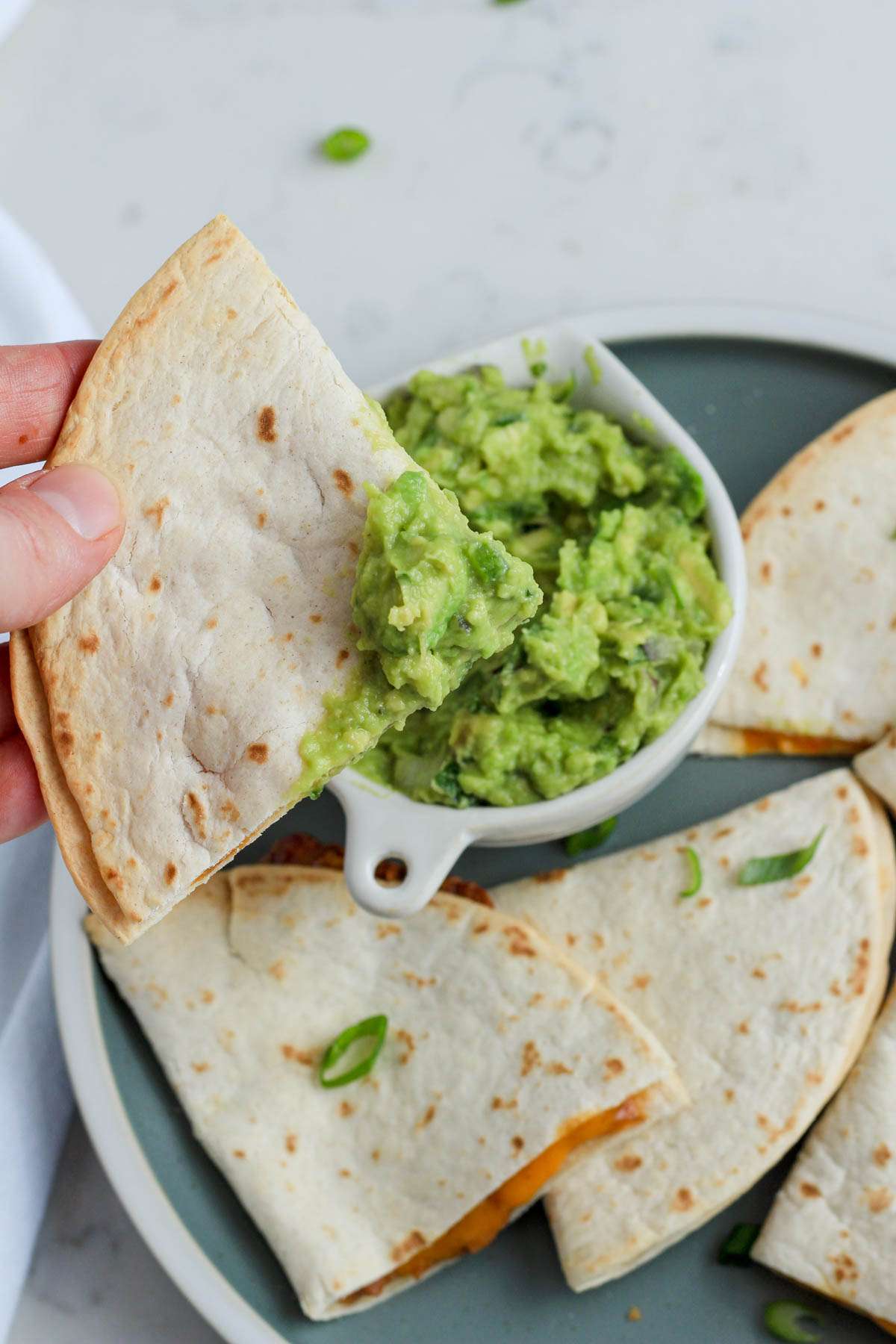 A hand holding a wedge of an air fryer quesadilla dipped in fresh guacamole.