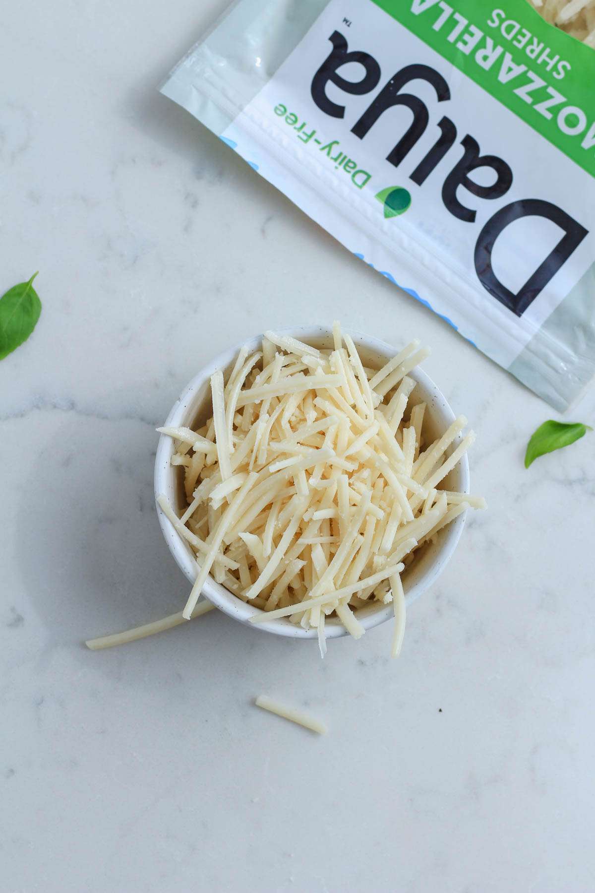 A small white bowl of Daiya mozzarella shreds on a white counter with a bag of the cheese in the top left corner.