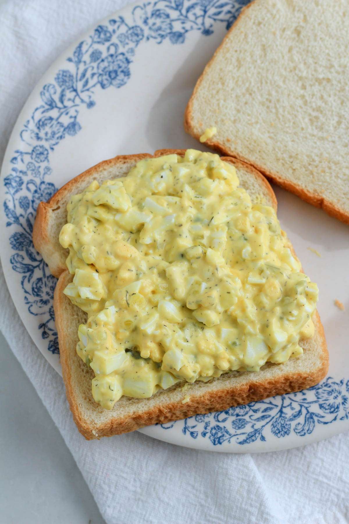 A blue and white plate on top of a white towel with a slice of bread topped with dill pickle egg salad.