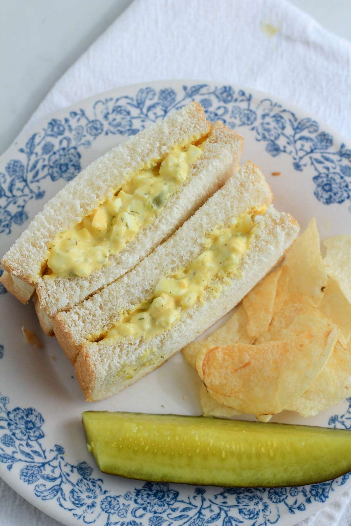 A white and blue plate with a dill pickle egg salad sandwich a small pile of potato chips and a dill pickle spear.