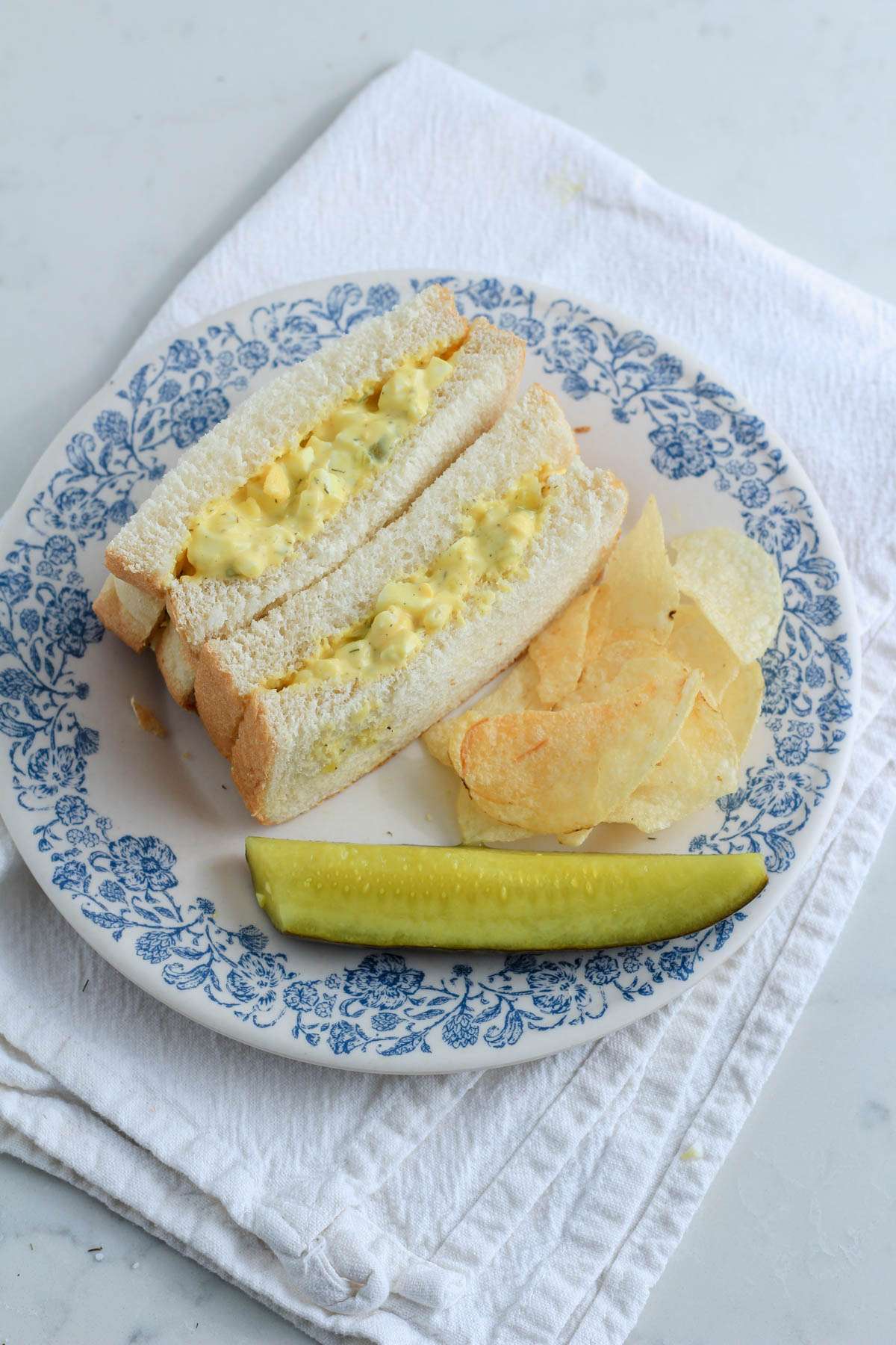 Dill pickle egg salad on a sandwich with a pickle and some potato chips on a white and blue plate.