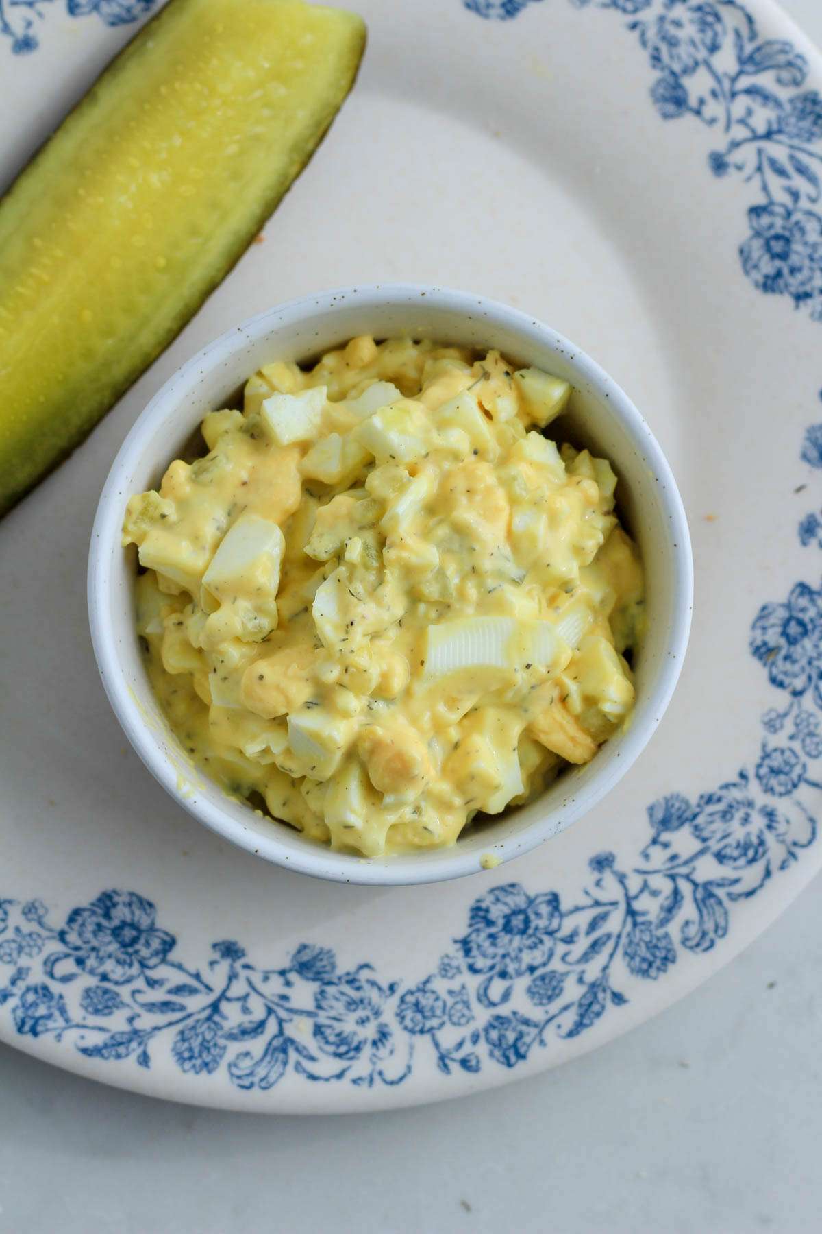 A top down picture of a small white bowl with dill pickle egg salad on a white and blue plate with a pickle spear to the left.