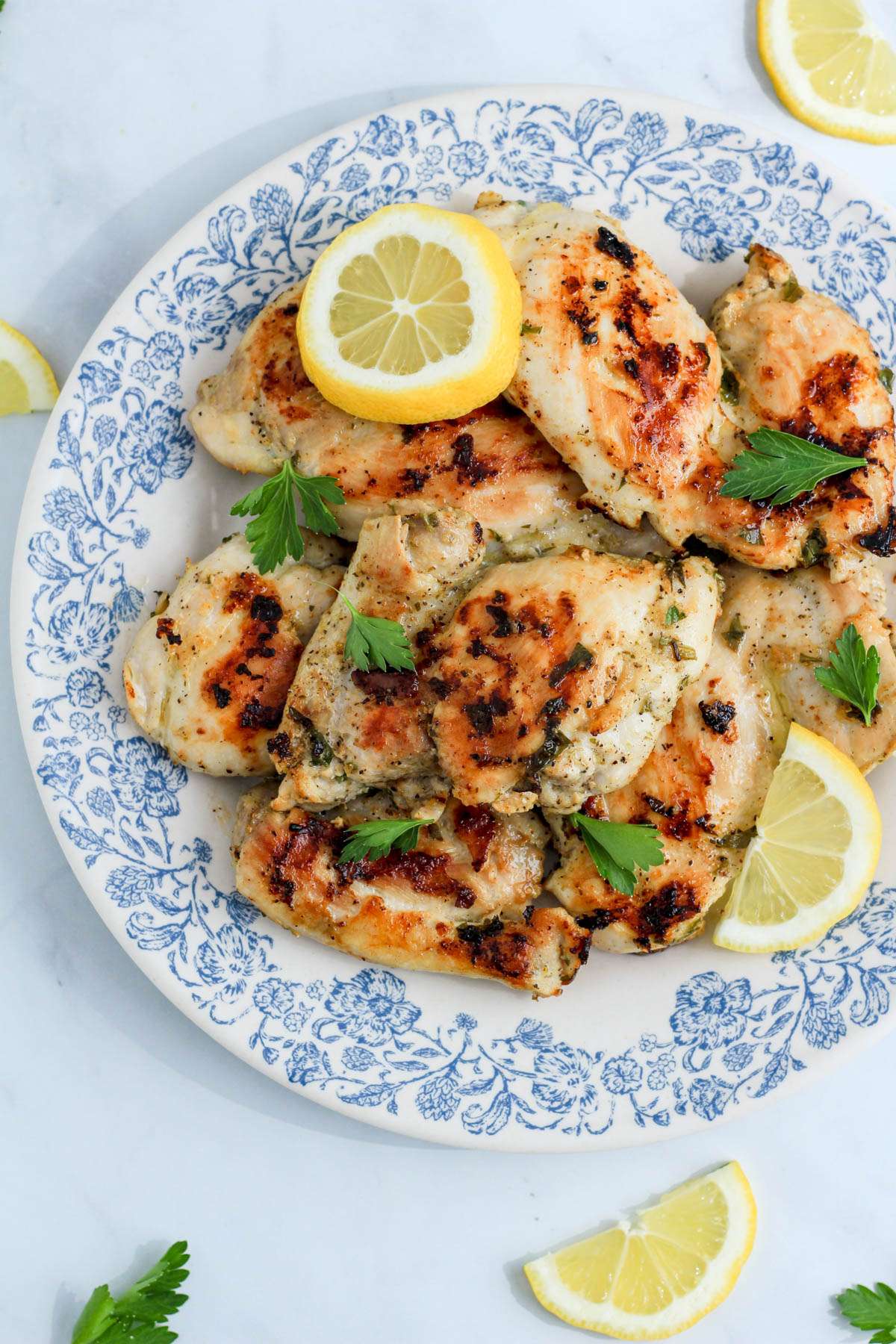 A white and blue bowl topped with Greek chicken thighs and lemon slices on a white counter.