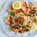A close up of a white and blue plate topped with grilled Greek marinated chicken thighs topped with parsley and lemon.
