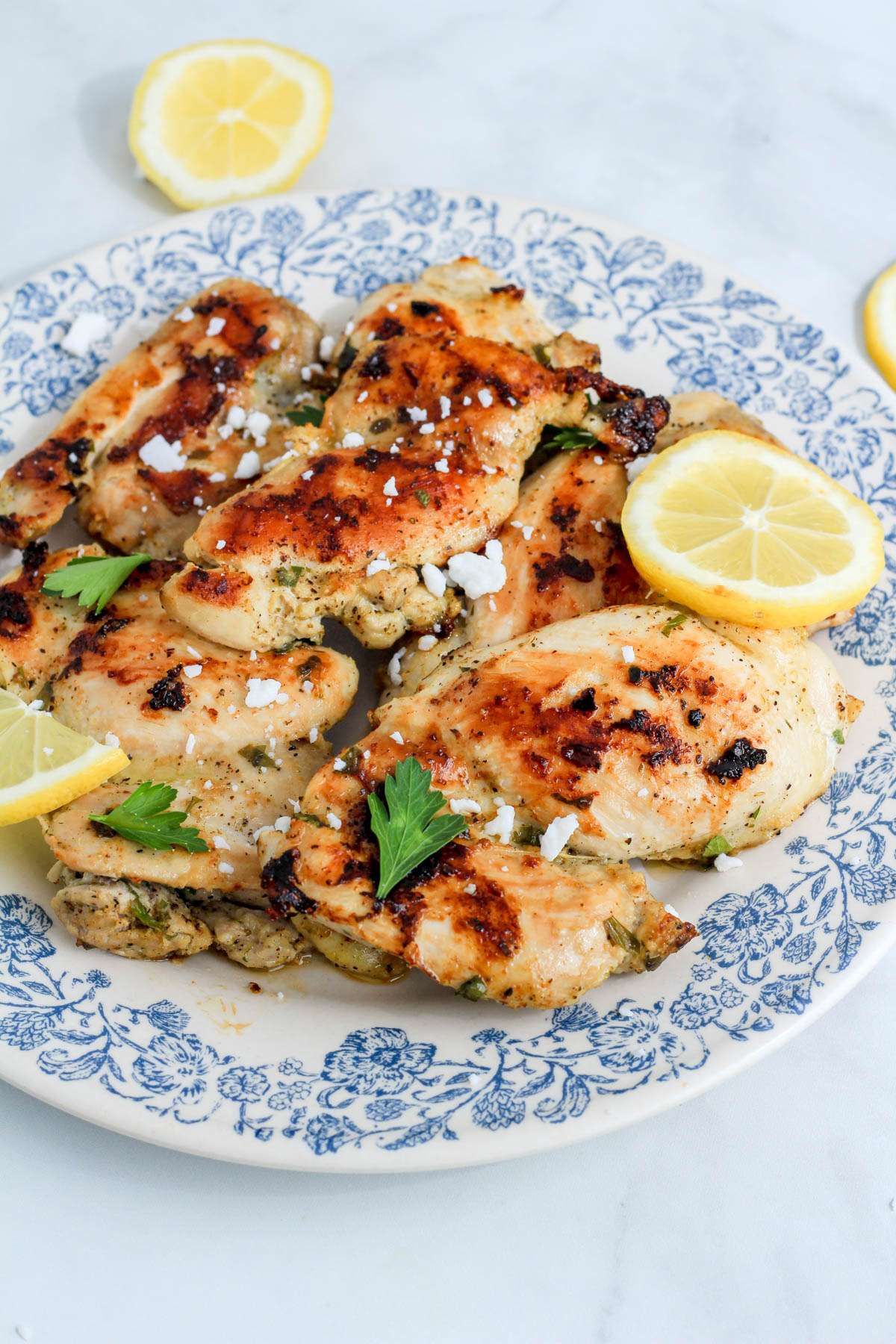 Vegan feta sprinkled on top of Greek marinated chicken thighs on a white and blue plate with lemon slices.