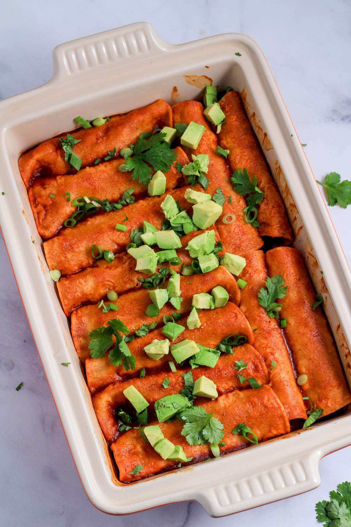 Cilantro and avocado topped ground turkey enchiladas in a rectangular pan on a white counter.